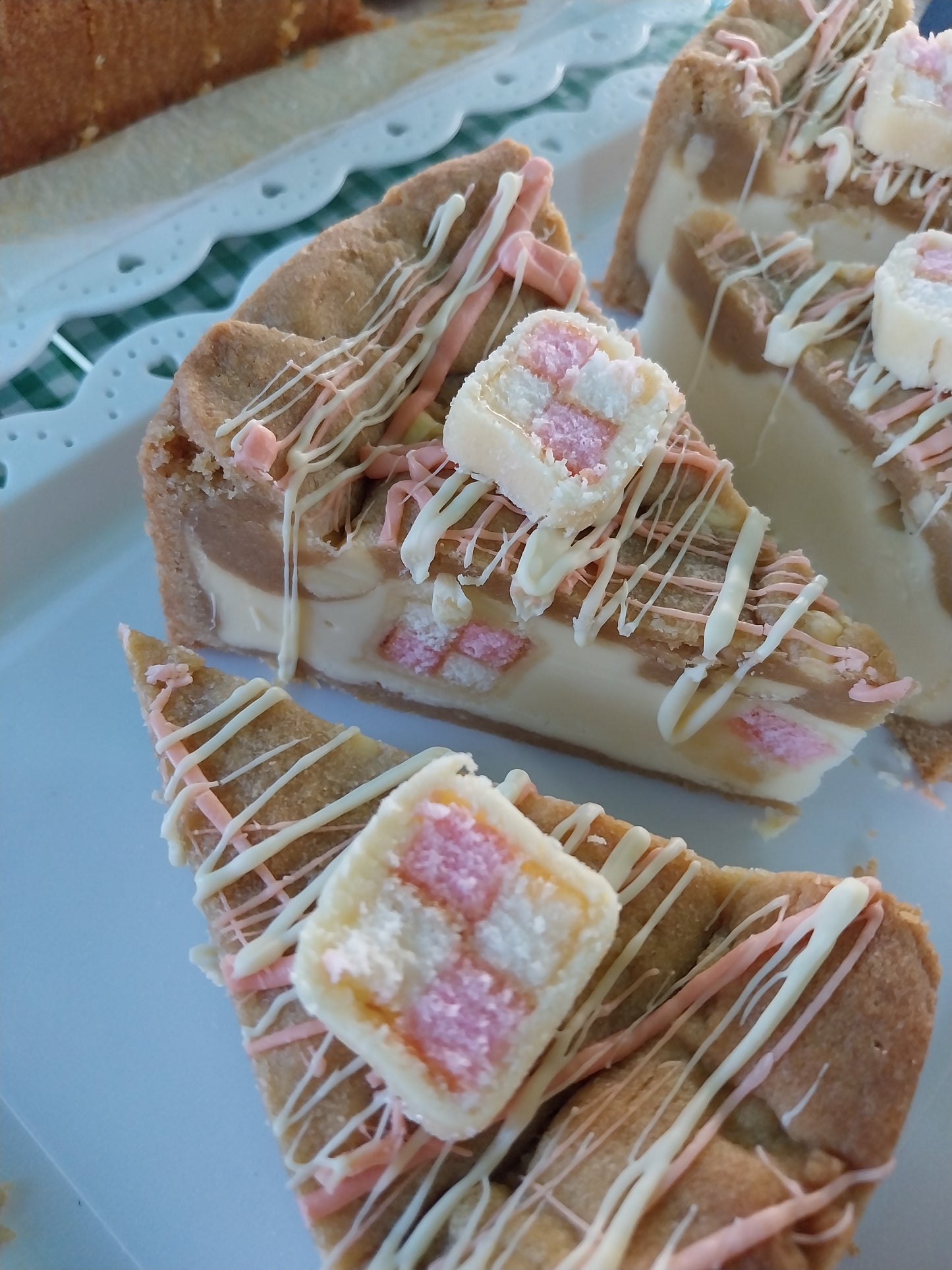 4 slices of Battenberg cookie pie on a tray. Filled with white chocolate spread and pieces of Battenberg cake and decorated with white and pink chocolate drizzle and a small slice of Battenberg cake on top.