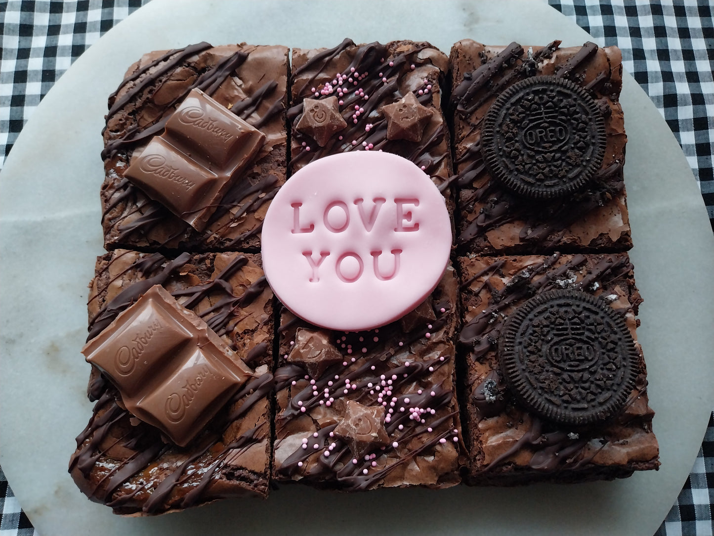 Six brownies on a marble slate. Two brownies are chocolate caramel with Cadbury caramel pieces. Two brownies are decorated with pink sprinkles and Magic Stars. Two brownies are topped with Oreos. Fondant topper says LOVE YOU