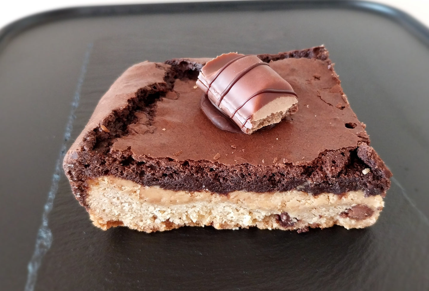 A slice of Kinder Bueno brookie on a dark slate. Kinder bueno brookie is made up of a layer of chocolate chip cookie, a layer of white chocolate hazelnut spread and chocolate brownie topped with a Kinder Bueno piece.