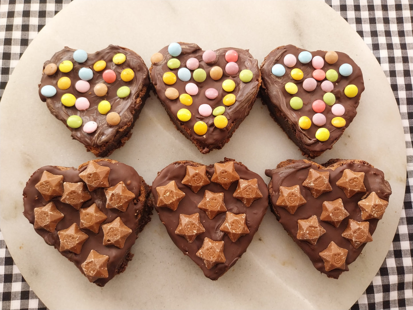 Six brownie hearts on a slate. All are decorated with a layer of dark chocolate. Three are topped with mini Smarties and three are topped with Magic Stars.