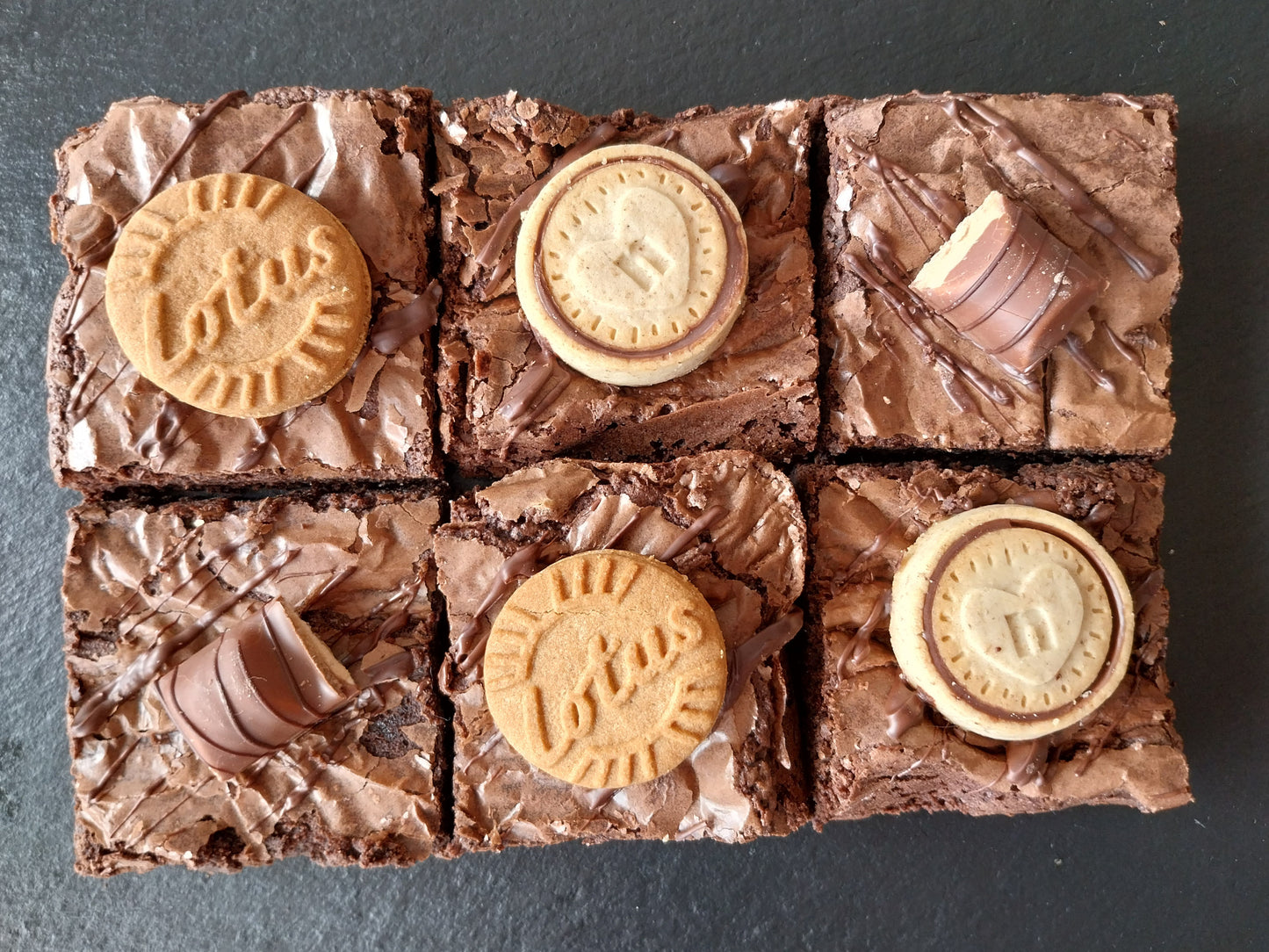 Six chocolate brownies on a slate. Two decorated with Biscoff biscuits, two decorated with Nutella biscuits and two with Kinder Bueno chunks