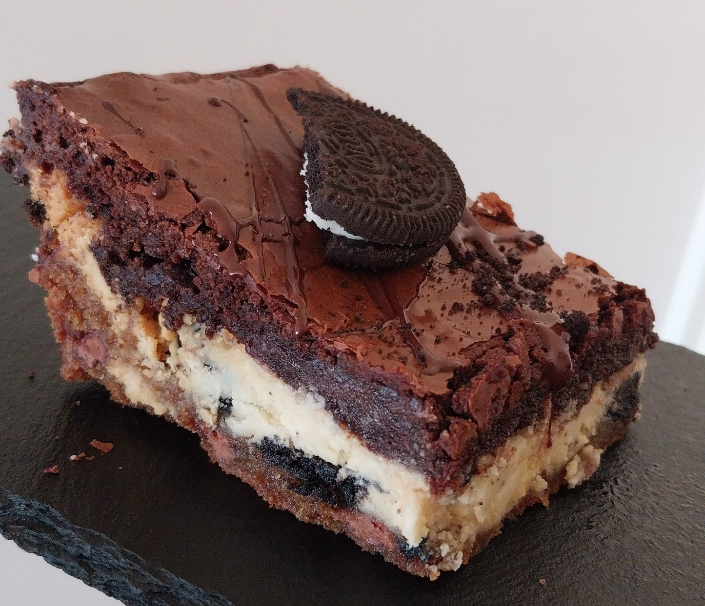 A slice of cookies & cream brookie on a dark slate. Cookies & cream brookie is made up of a layer of chocolate chip cookie, a layer of white chocolate spread with Oreos and chocolate brownie topped with an Oreo.