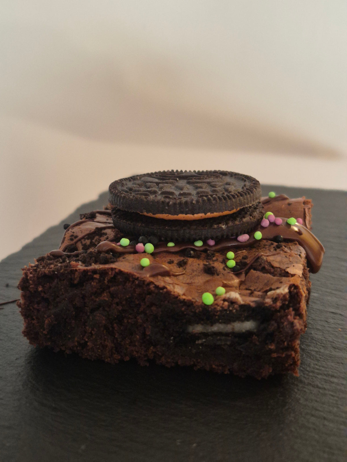 A chocolate brownie slice decorated with a whole spooky Oreo cookie and colorful sprinkles, shown on a dark slate surface.
