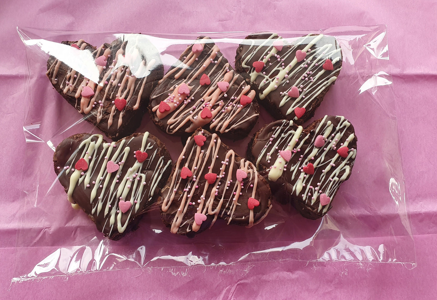 6 heart shaped brownies decorated with a layer of dark chocolate, drizzled with white chocolate and heart sprinkles. Wrapped in a clear cellophane bag on pink tissue paper