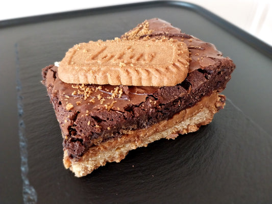 One slice of Biscoff brookie on a dark slate. Layer of cookie, Biscoff spread and chocolate brownie topped with Biscoff biscuit.