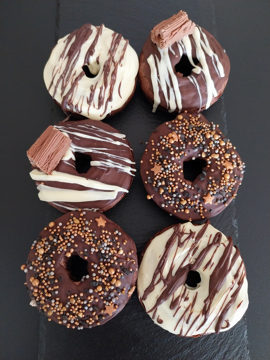 Six chocolate bronuts on a dark slate. Two bronuts are decorated with white chocolate and milk chocolate drizzle, two decorated with dark chocolate and white chocolate drizzle and a Cadbury Flake. Two bronuts have sprinkles on top.