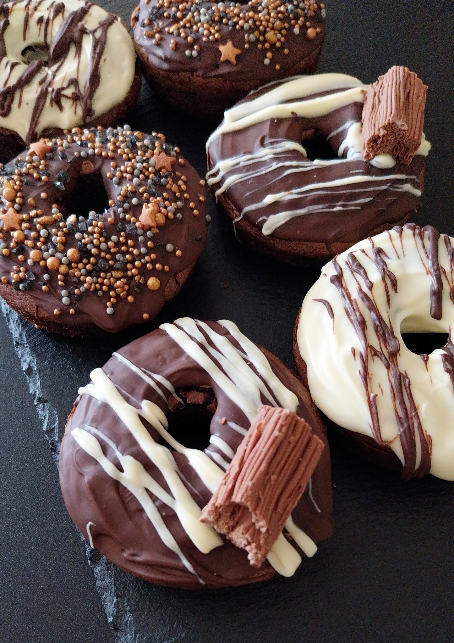 Six chocolate bronuts on a dark slate. Two bronuts are decorated with white chocolate and milk chocolate drizzle, two decorated with dark chocolate and white chocolate drizzle and a Cadbury Flake. Two bronuts have sprinkles on top.