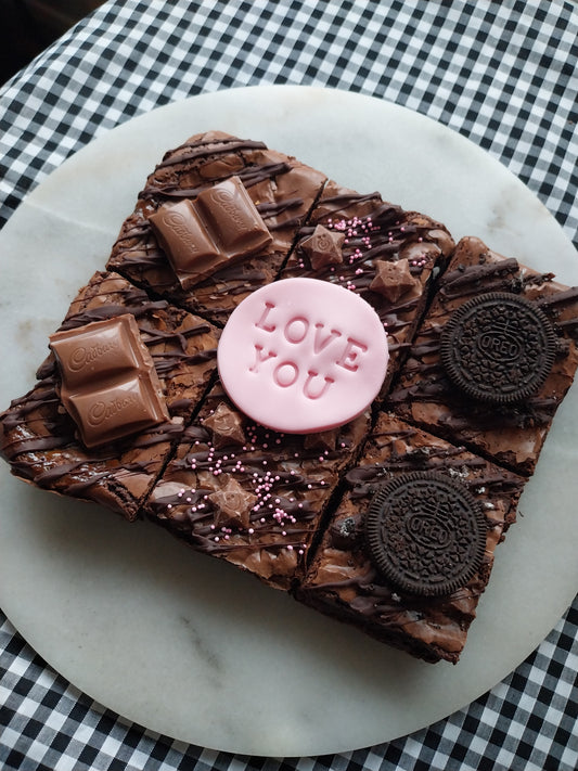 Six brownies on a marble slate. Two brownies are chocolate caramel with Cadbury caramel pieces. Two brownies are decorated with pink sprinkles and Magic Stars. Two brownies are topped with Oreos. Fondant topper says LOVE YOU