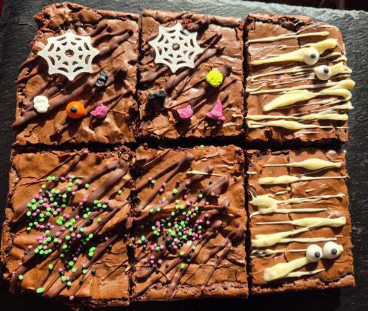 Six chocolate brownies. 4 are decorated with halloween themed sprinkles and cobweb. 2 are decorated with white chocolate drizzle and 2 edible eyes in the style of a mummy