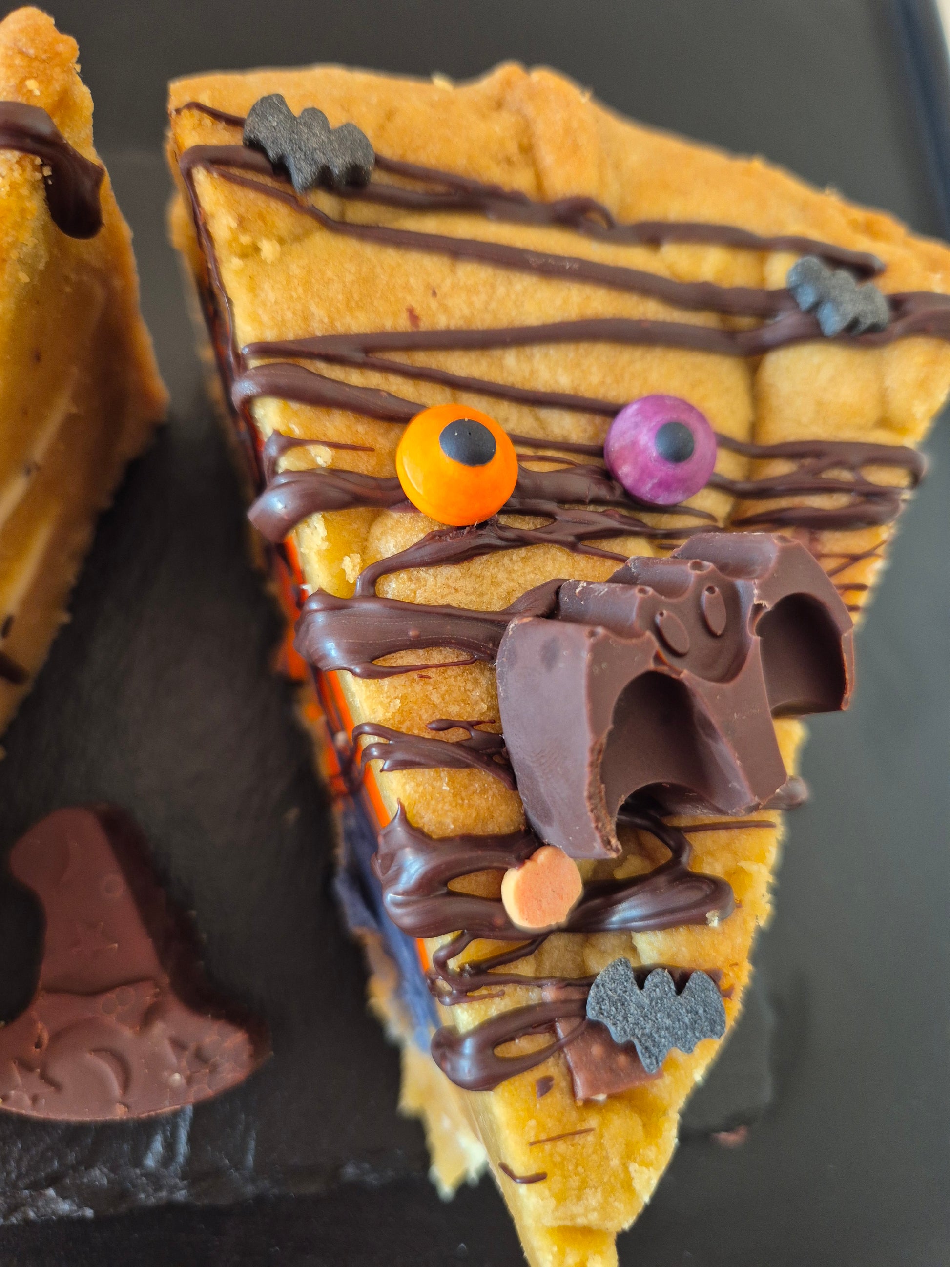 Halloween-themed cookie pie slice with chocolate drizzle, candy eyes, and decorative elements on a dark surface.