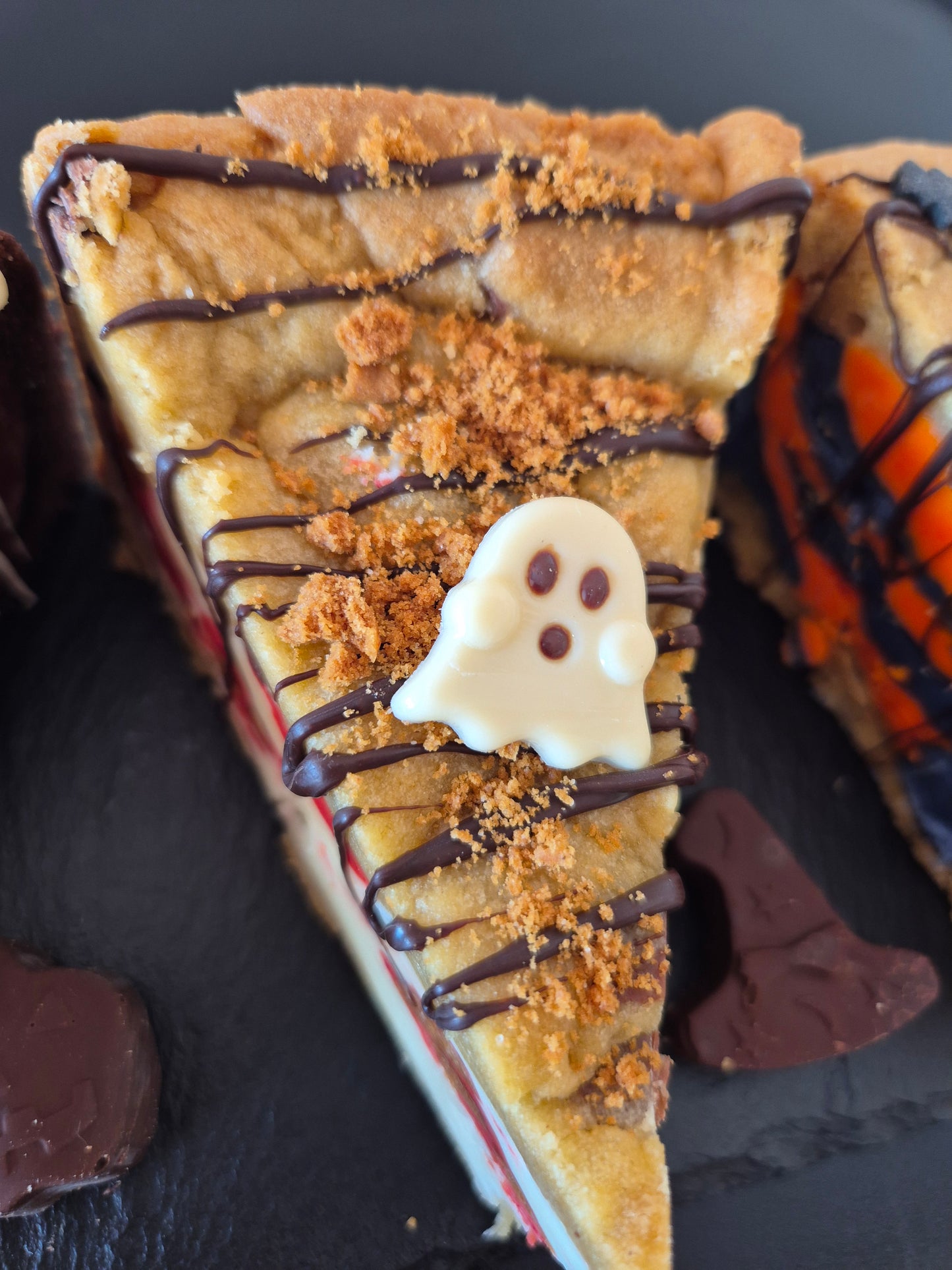 Halloween-themed cookie pie with a ghost face made of white chcocolate and chocolate drizzle on a dark surface.