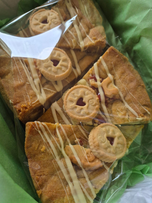 Box of 4 jammie dodger cookie pie slices wrapped in cellophane on green tissue paper. Each cookie pie slice is topped with white chocolate drizzle and mini jammie dodger biscuits