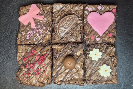 Six chocolate brownies on a dark slate topped with different decorations including chocolate orange segment, Lindor chocolate, white chocolate flowers, dried raspberry pieces, pink fondant heart and bow and pink and heart sprinkles
