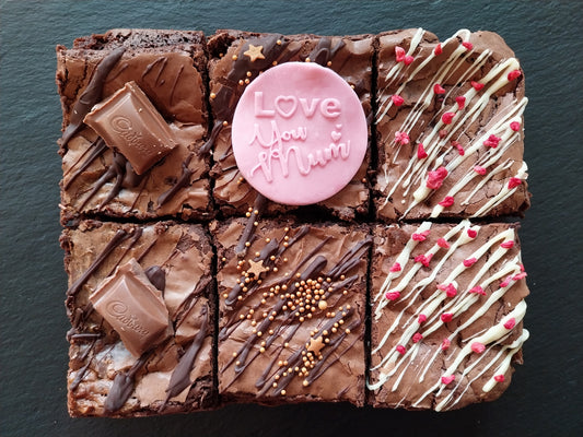 Six chocolate brownies. Two are topped with Cadbury Caramel chocolates. One is topped with a pink fondant topper which says 'Love you mum'. One is drizzled with dark chocolate and orange sprinkles. Two are decorated with white chocolate and raspberry