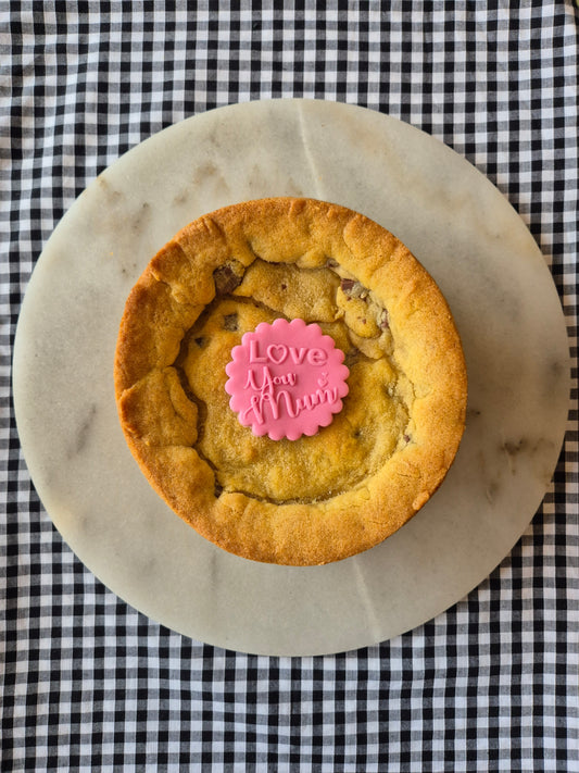 Whole cookie pie on a marble slate on black and white gingham. The cookie pie has a pink fondant topper with the words Love You Mum on it