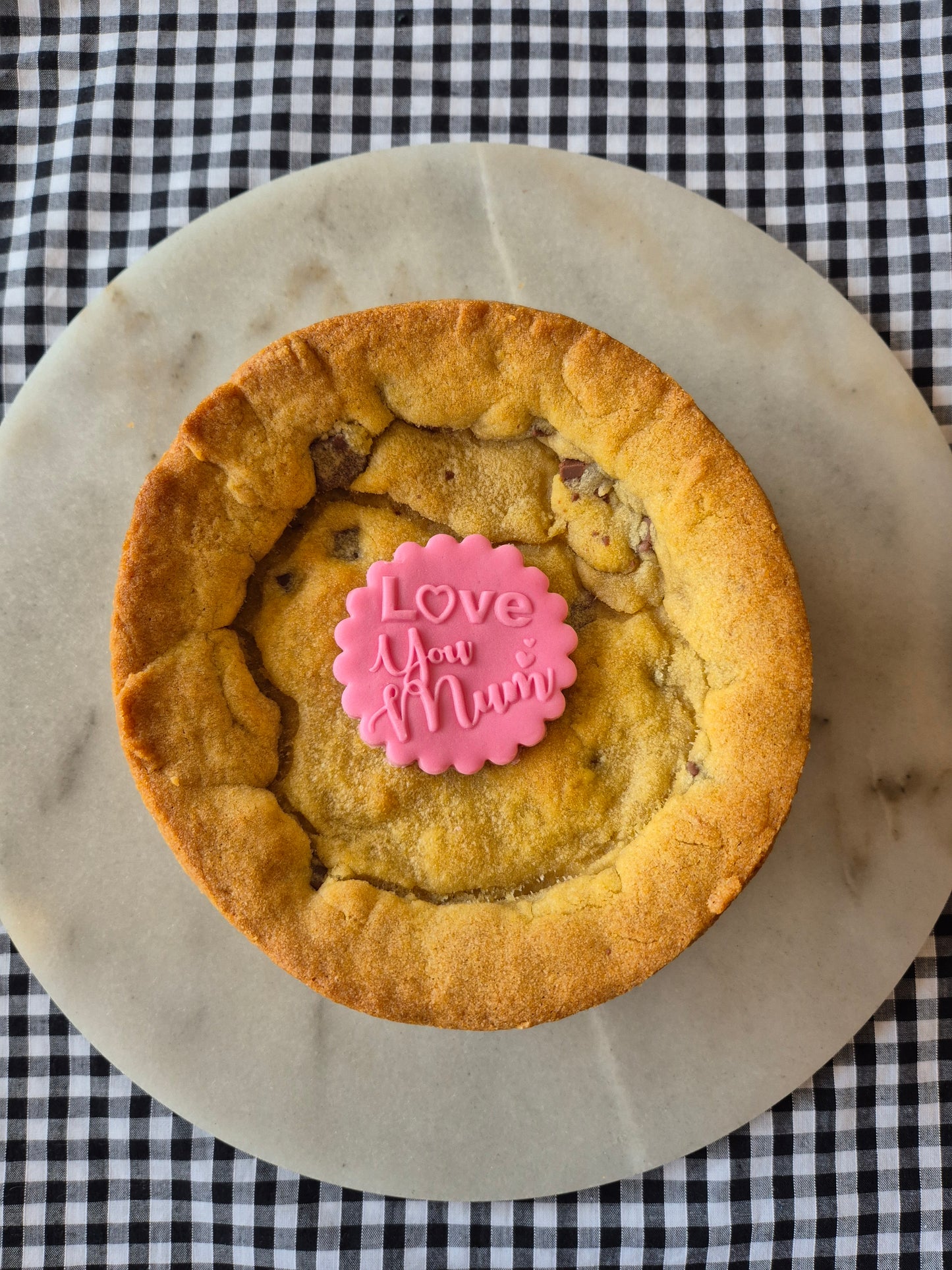 Whole cookie pie on a marble slate on black and white gingham. The cookie pie has a pink fondant topper with the words Love You Mum on it