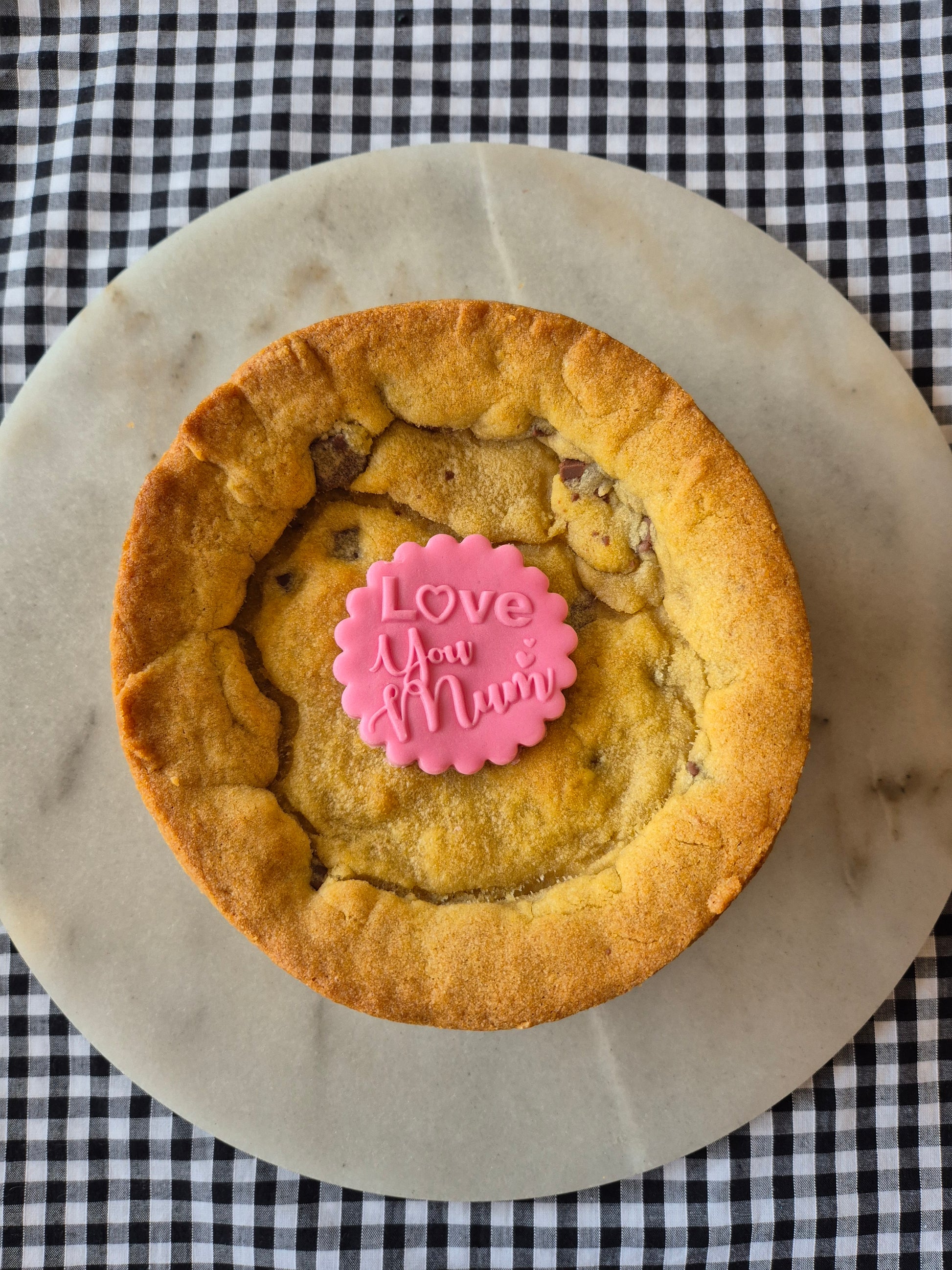 Whole cookie pie on a marble slate on black and white gingham. The cookie pie has a pink fondant topper with the words Love You Mum on it