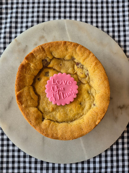 Whole cookie pie on a marble slate on black and white gingham. The cookie pie has a pink fondant topper with the words Love You Mum on it