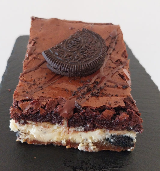 A slice of cookies & cream brookie on a dark slate. Cookies & cream brookie is made up of a  layer of chocolate chip cookie, a layer of white chocolate spread with Oreos and chocolate brownie topped with an Oreo.