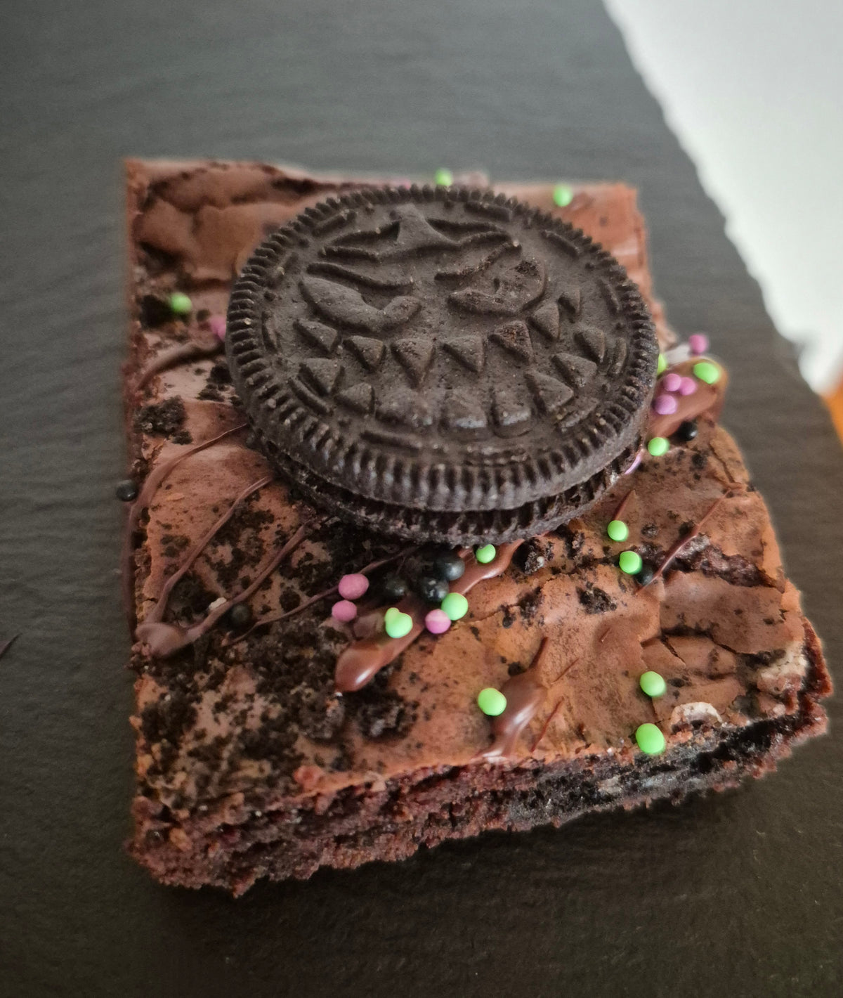 A chocolate brownie topped with a spooky Oreo cookie, drizzled with chocolate, and sprinkled with colorful candy-coated sprinkles, presented on a dark slate surface.