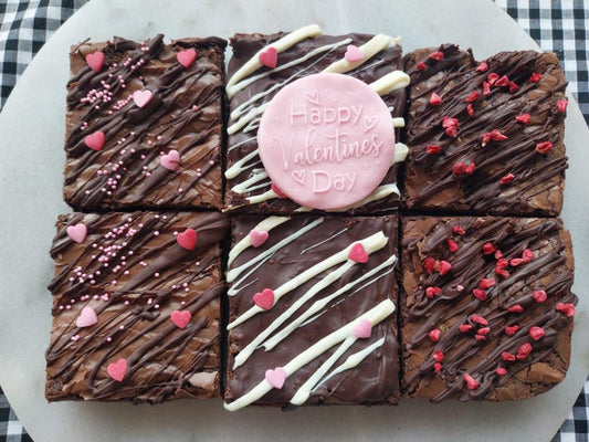 Six chocolate brownies all decorated with chocolate drizzle, sprinkles and one brownie has a pink fonder topper on it which says 'Happy Valentines Day'. 