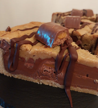 A close-up image of a Wispa Gold Cookie Pie slice showing the chocolate chip cookie base, a layer of milk chocolate spread, and chunks of Wispa Gold chocolate bars with a dark chocolate drizzle on top.