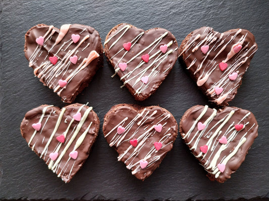 6 heart shaped brownies decorated with a layer of dark chocolate, drizzled with white chocolate and heart sprinkles. On a slate background